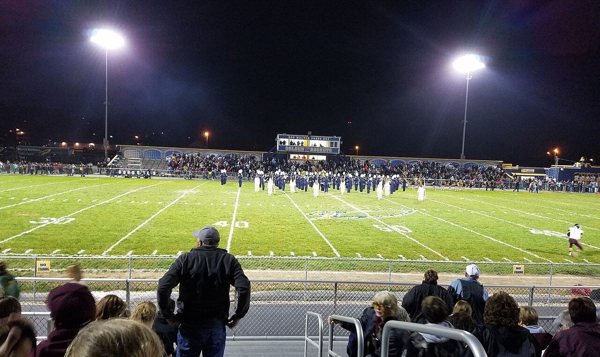 A Friday night high school football game under floodlights with a full crowd